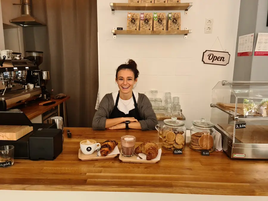 Barista lächelt hinter der Holztheke, davor Kaffee, Croissants und Kekse