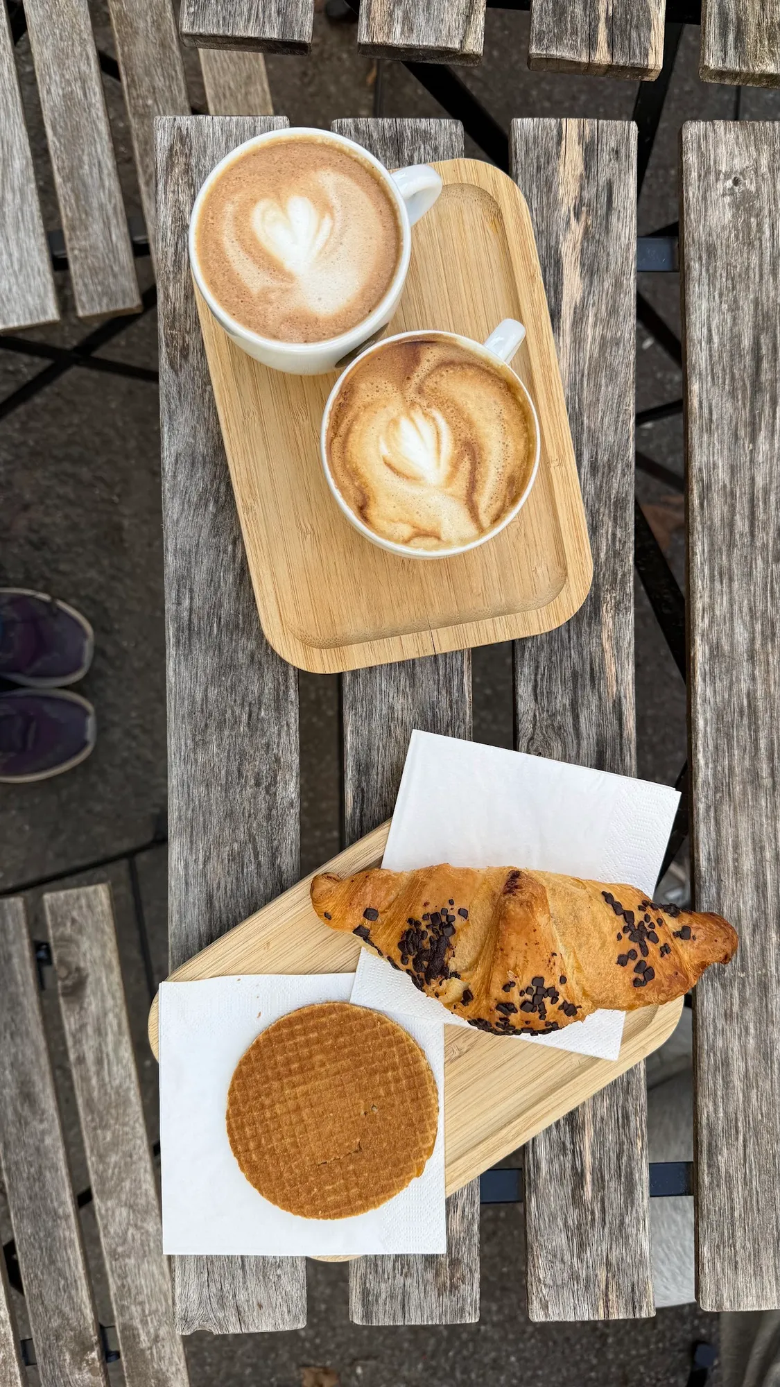 Zwei Cappuccinos, ein Croissant und eine Waffel auf einem Holztisch im Freien